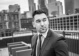 A man in a suit poses on a rooftop with a city skyline.